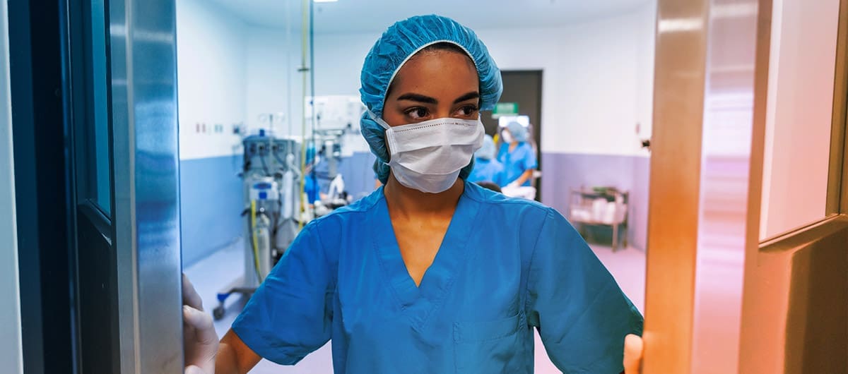 Healthcare worker in scrubs, mask, and hairnet captured walking out of a care room while pushing open swinging doors