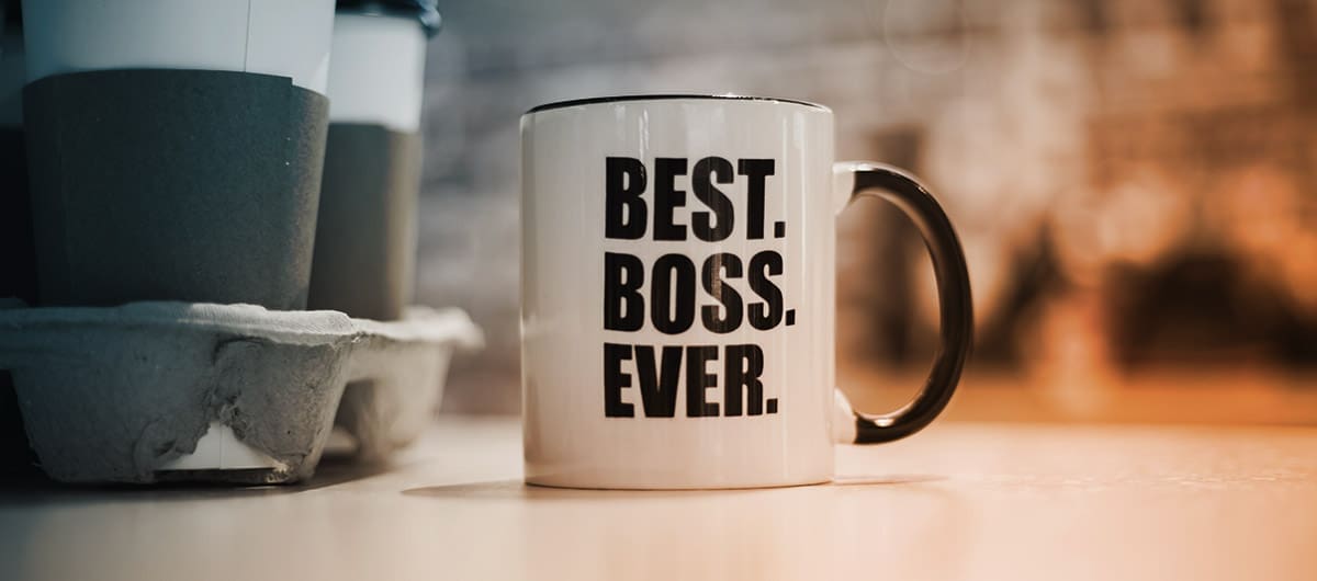 A white mug with black text reading "Best. Boss. Ever." next to a drink carrier holding to-go coffee cups, sitting on a counter