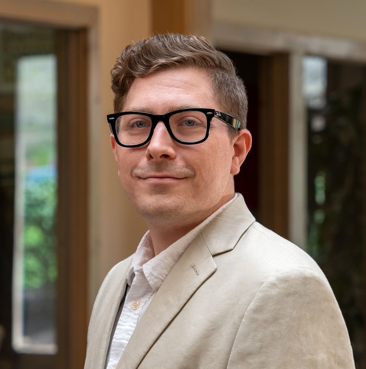 Headshot of a grinning man with brown, wavy hair dressed in a blazer and glasses in front of a blurred background