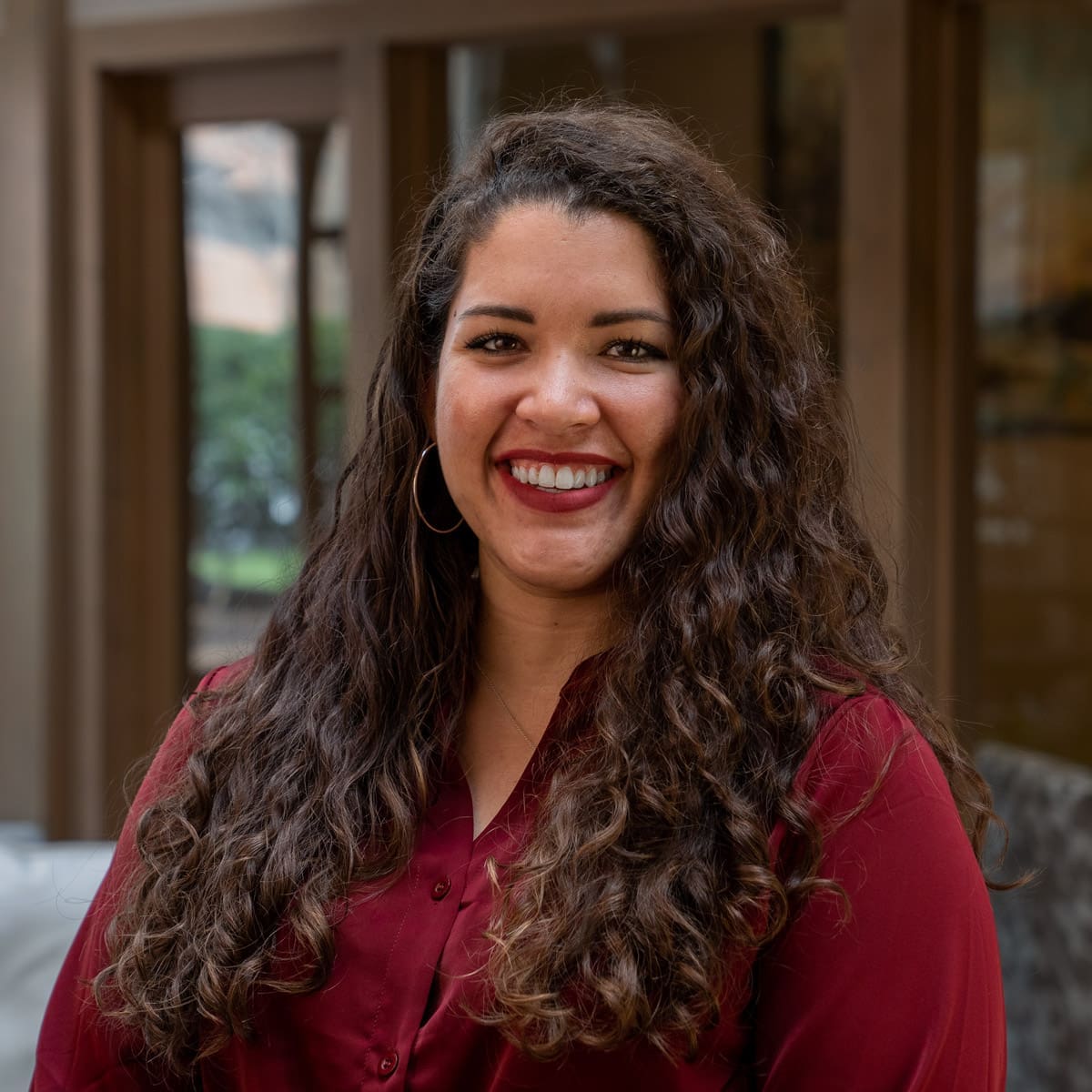 Headshot of a smiling woman with curly, dark brown hair dressed in corporate attire in front of a blurred background