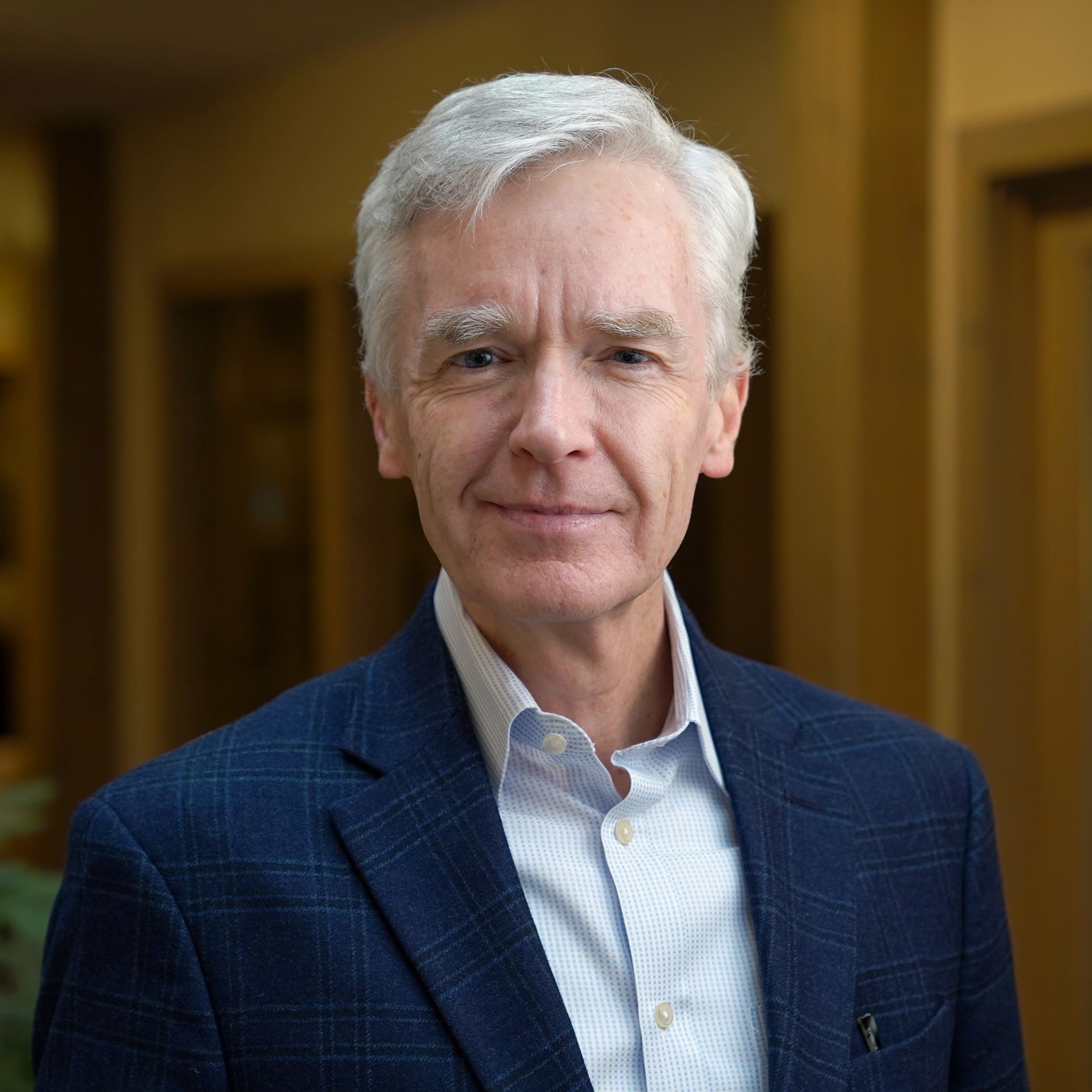 Headshot of a grinning man with white hair dressed in a blazer in front of a blurred background