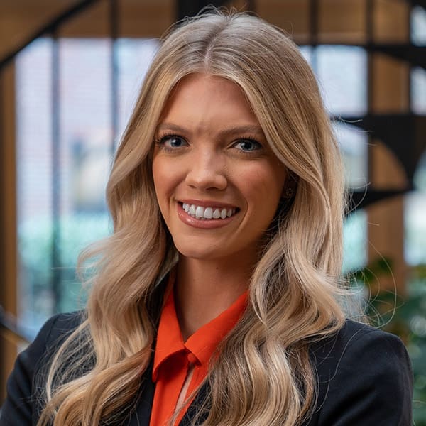 Headshot of a smiling young woman with blonde hair dressed in a blazer in front of a blurred background