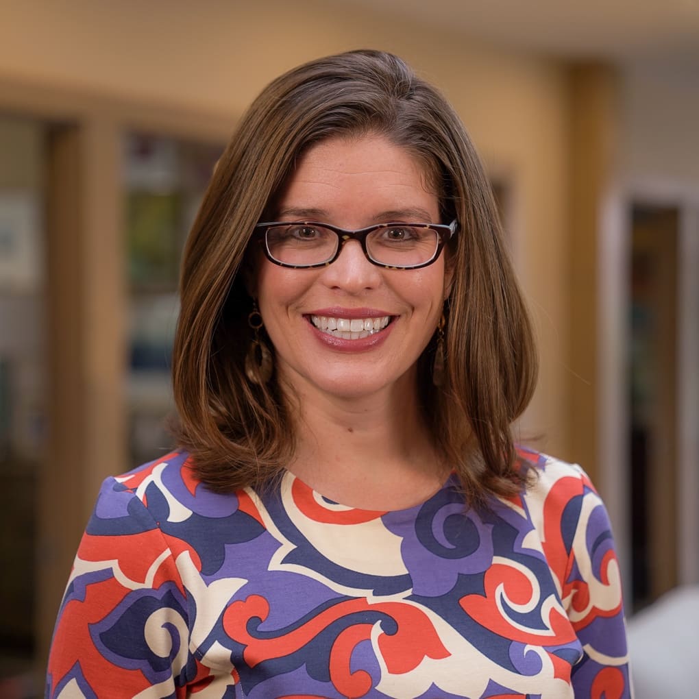 Headshot of a smiling woman with short, brunette hair and glasses, dressed in corporate attire in front of a blurred background