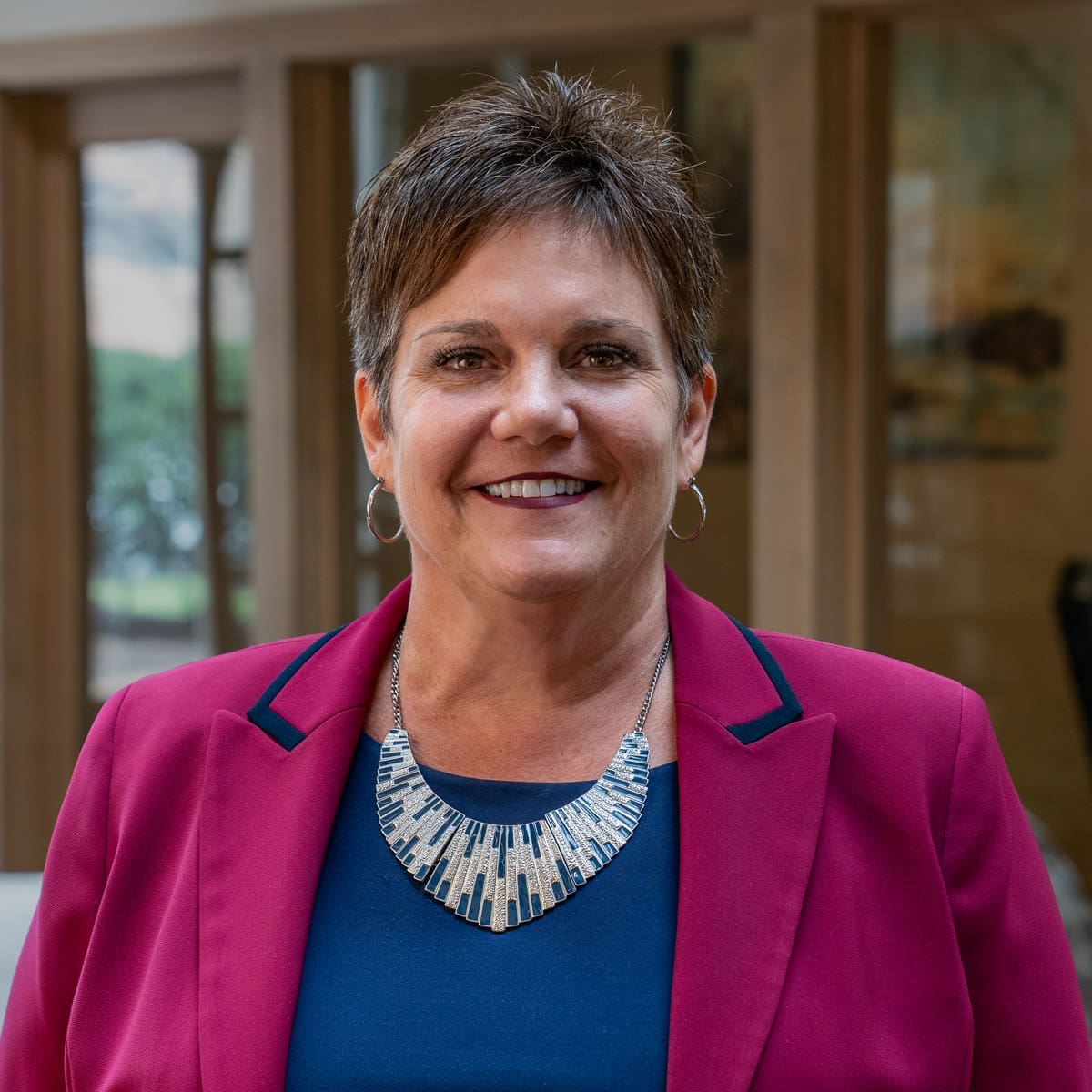 Headshot of a smiling woman with short, brunette hair dressed in a pink blazer in front of a blurred background