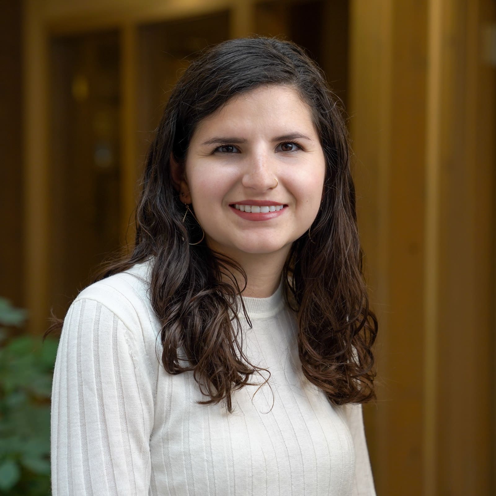 Headshot of a smiling young woman with dark brunette hair dressed in a white sweater in front of a blurred background