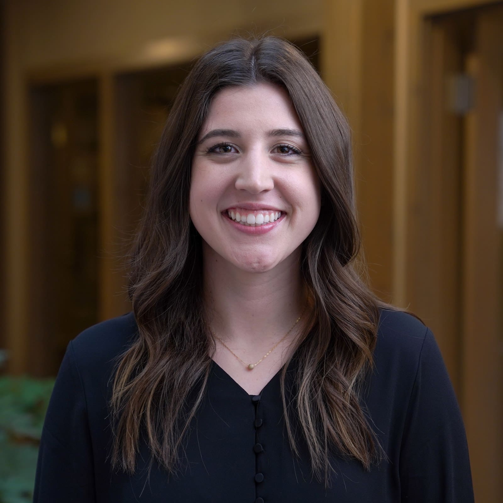 Headshot of a smiling young woman with dark brunette hair dressed in corporate attire in front of a blurred background