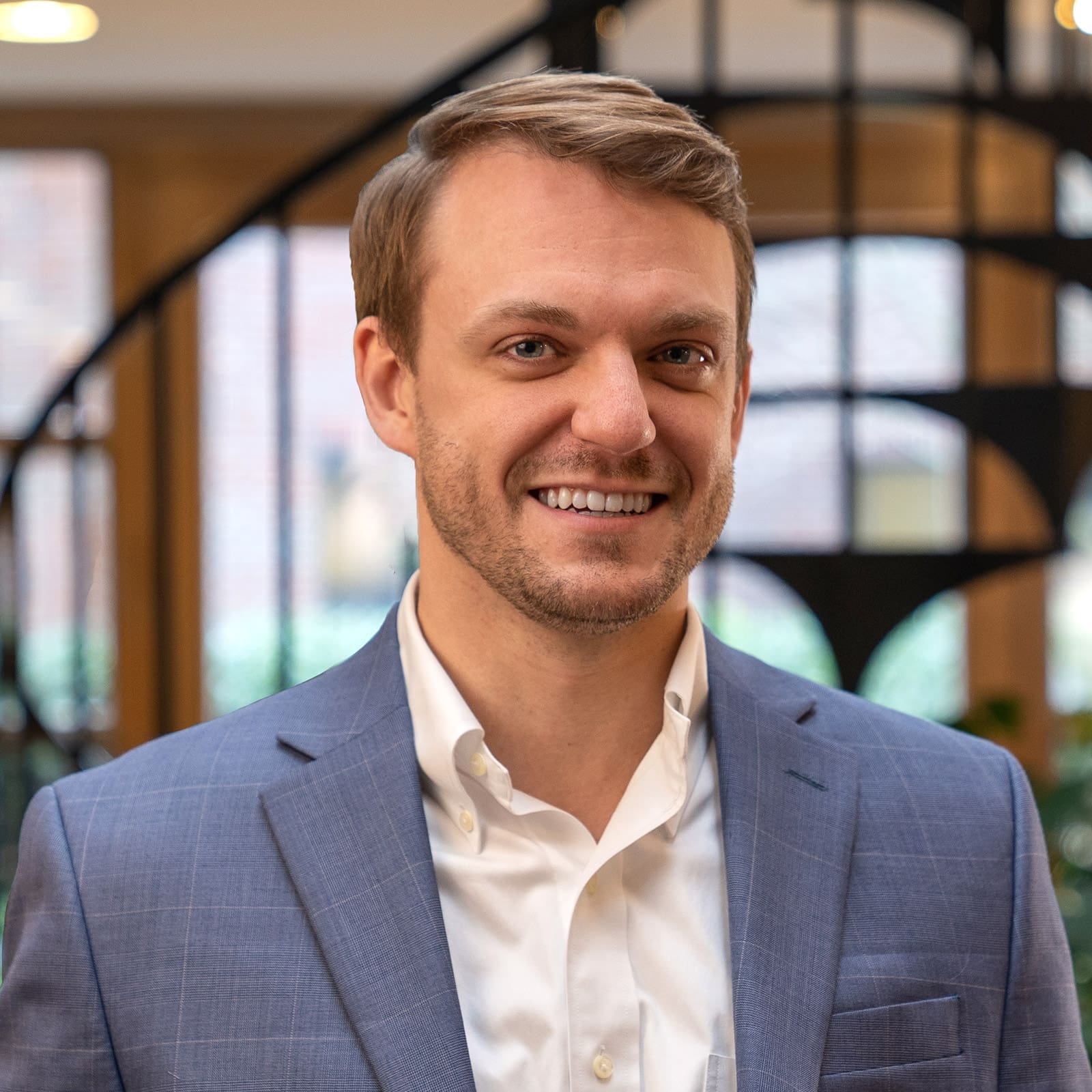 Headshot of a smiling young man with blonde hair dressed in a blazer in front of a blurred background