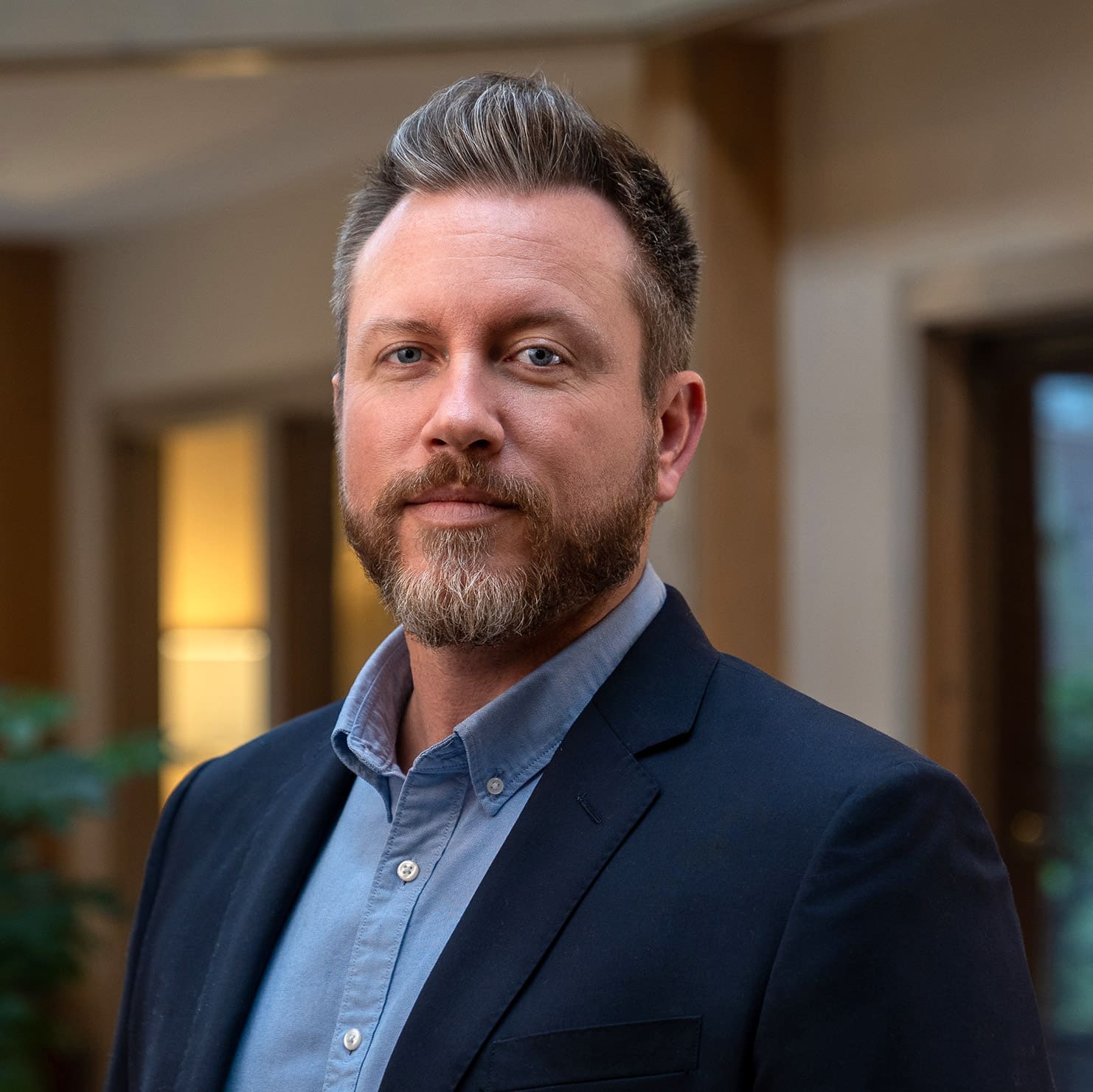 Headshot of a grinning man with light brown hair and beard, dressed in a blazer in front of a blurred background