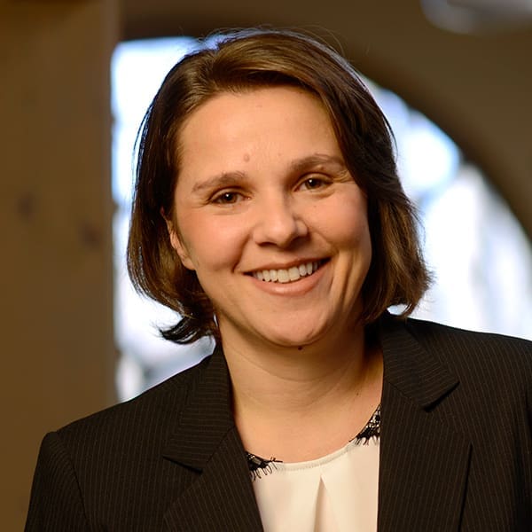 Headshot of a smiling woman with short, brunette hair dressed in a blazer in front of a blurred background