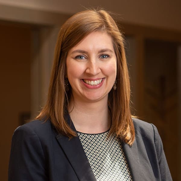 Headshot of a smiling woman with reddish-brown hair dressed in a blazer in front of a blurred background