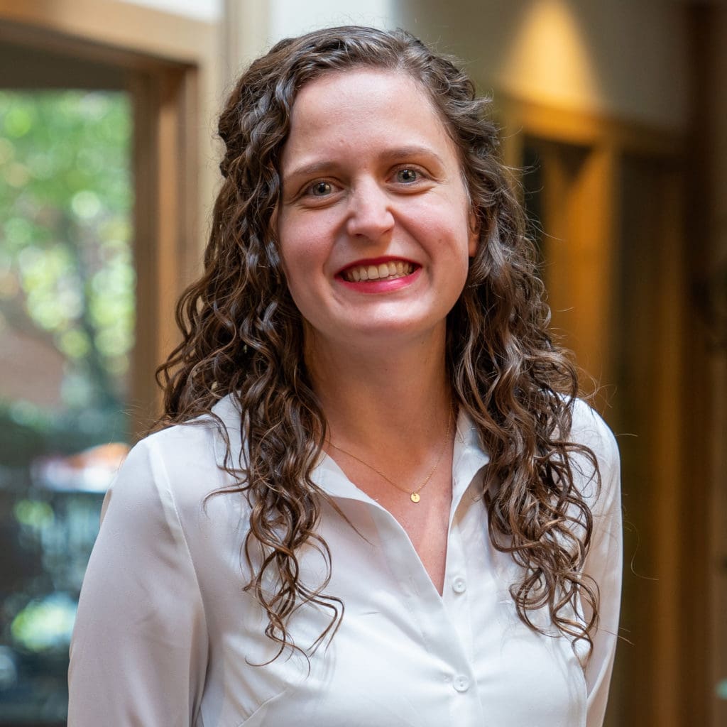 Headshot of a smiling young woman with brunette, curly hair dressed in corporate attire in front of a blurred background